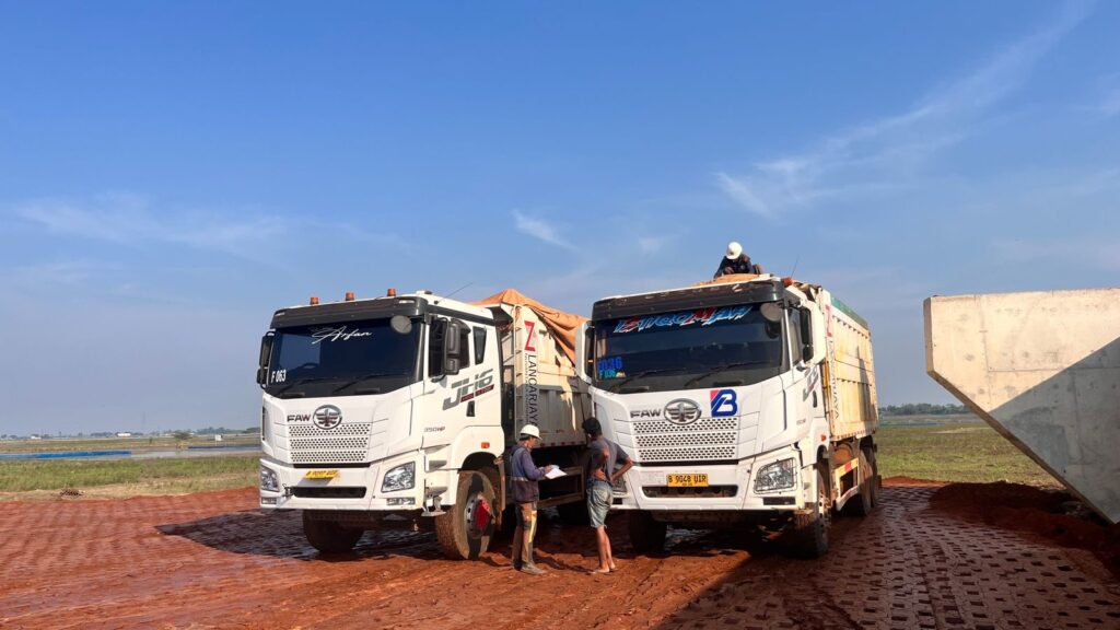 Dua unit dump truck dari PT Berkat Bumi Persada (BBSP) sedang beroperasi di lokasi proyek konstruksi jalan Tol Akses Patimban, dengan latar belakang langit biru cerah dan area pembangunan.