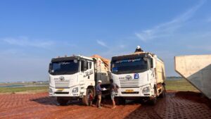 Dua unit dump truck dari PT Berkat Bumi Persada (BBSP) sedang beroperasi di lokasi proyek konstruksi jalan Tol Akses Patimban, dengan latar belakang langit biru cerah dan area pembangunan.