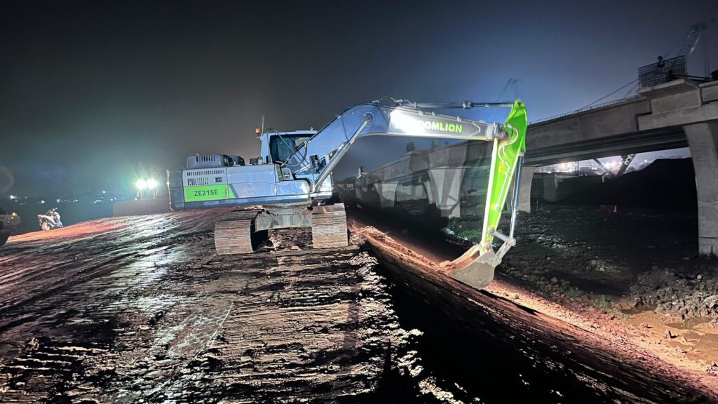 Ekskavator besar milik PT BBSP dengan lampu sorot sedang meratakan tanah di malam hari pada pembangunan Proyek Tol Akses Patimban. Terlihat struktur jalan tol yang sedang dalam tahap konstruksi di latar belakang